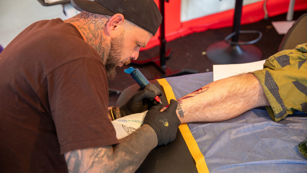 Tätowierer sticht einem Festivalbesucher beim Göss OpenAir 2026 in Gössmannsreuth ein Tattoo auf den Unterarm.